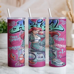 Pu&ograve; includere: Un bicchiere cilindrico con sfondo rosa raffigurante un personaggio dei cartoni animati con capelli rossi, un granchio e un boombox. Il design include scritte in stile graffiti Ariel, Free Spirit, Baddie, Rebel e Goddess, con una cannuccia bianca.