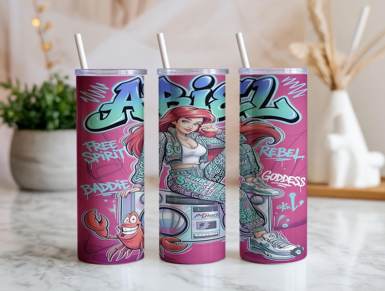 Pu&ograve; includere: Un bicchiere cilindrico con sfondo rosa raffigurante un personaggio dei cartoni animati con capelli rossi, un granchio e un boombox. Il design include scritte in stile graffiti Ariel, Free Spirit, Baddie, Rebel e Goddess, con una cannuccia bianca.