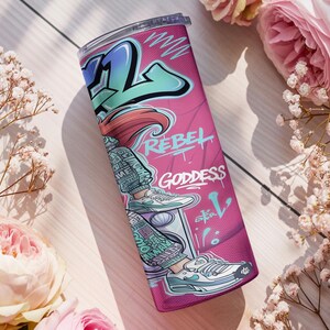 Pu&ograve; includere: Un bicchiere cilindrico con sfondo rosa e grafica in stile graffiti. Il design include una figura con scarpe da ginnastica, la scritta REBEL e GODDESS e motivi astratti blu e bianchi. Il bicchiere &egrave; dotato di un coperchio trasparente.
