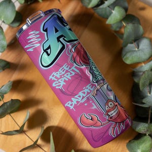 Pu&ograve; includere: Un bicchiere cilindrico in acciaio inossidabile con sfondo rosa, caratterizzato da una grande lettera A in stile graffiti, un granchio dei cartoni animati e il testo "FREE SPIRIT" e "BADDIE" in un carattere stilizzato. Include un coperchio trasparente.