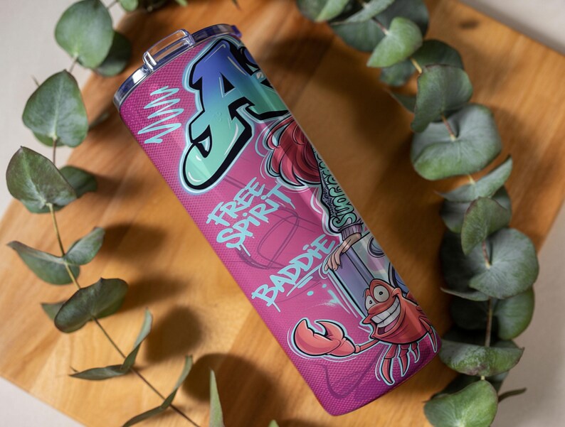 Pu&ograve; includere: Un bicchiere cilindrico in acciaio inossidabile con sfondo rosa, caratterizzato da una grande lettera A in stile graffiti, un granchio dei cartoni animati e il testo "FREE SPIRIT" e "BADDIE" in un carattere stilizzato. Include un coperchio trasparente.