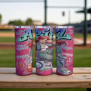 Pu&ograve; includere: Tre bicchieri cilindrici con sfondo rosa raffiguranti un personaggio dei cartoni animati con capelli rossi, un granchio e un boombox. Il design include scritte in stile graffiti che recitano "ARIEL", "FREE SPIRIT", "BADDIE", "REBEL" e "GODDESS".
