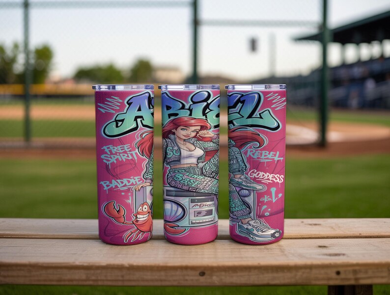 Pu&ograve; includere: Tre bicchieri cilindrici con sfondo rosa raffiguranti un personaggio dei cartoni animati con capelli rossi, un granchio e un boombox. Il design include scritte in stile graffiti che recitano "ARIEL", "FREE SPIRIT", "BADDIE", "REBEL" e "GODDESS".