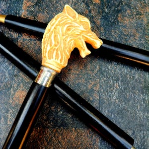 May include: A black walking stick featuring a carved wolf head handle in cream and tan hues. The handle is connected to the black shaft with a silver-toned metal band. The stick rests on a dark, textured surface.