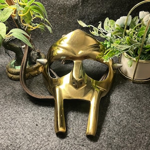 Può includere: Una maschera in metallo color oro dal design distintivo. La maschera presenta un elemento nasale prominente, due aperture per gli occhi e due punte sui lati. È attaccato un cinturino marrone. È un oggetto decorativo.