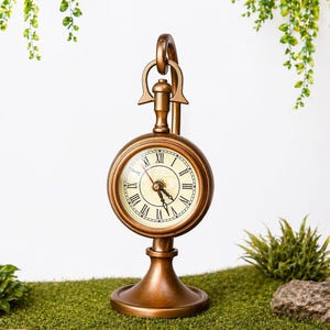 Puede incluir: Un reloj de escritorio de estilo vintage, de color bronce, con esfera redonda y números romanos. El reloj tiene un asa decorativa y se apoya sobre una base. El reloj se exhibe sobre musgo verde.