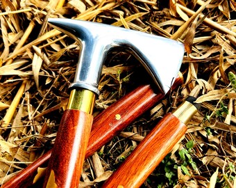 May include: A silver-colored metal walking cane with a curved handle and a polished wooden shaft. The shaft is a rich, reddish-brown color with brass accents. The cane is resting on a bed of dried leaves.