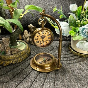 May include: A vintage brass pocket watch and compass set. The pocket watch has Roman numerals and the text "Victoria London". The compass is below the watch, and the set is on a decorative stand. The background includes greenery and flowers.