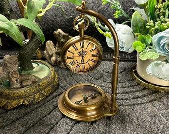 May include: A vintage brass pocket watch and compass set. The pocket watch has Roman numerals and the text "Victoria London". The compass is below the watch, and the set is on a decorative stand. The background includes greenery and flowers.