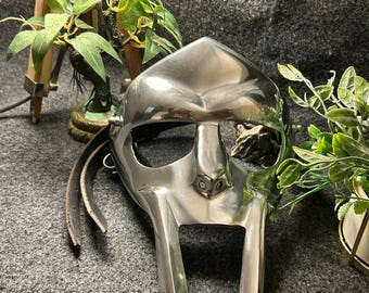 May include: A polished silver metal mask with a distinctive, angular design. The mask features a prominent nose piece and eye openings, with leather straps attached. The overall aesthetic suggests a futuristic or stylized helmet.