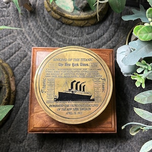 Może przedstawiać: Okrągła, mosiężna plakietka z nagłówkiem "Sinking of the Titanic" z The New York Times, z 16 kwietnia 1912 roku. Plakietka jest zamontowana na kwadratowej, drewnianej podstawie. Obraz przedstawia ilustrację Titanica.