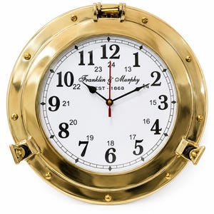 May include: A round wall clock with a polished brass porthole frame, featuring a white face with black Arabic numerals from 1 to 12 and smaller 13 to 24 hour markers. The centre displays the text Franklin & Murphy EST 1868 with black hands.