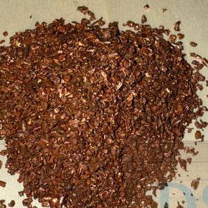 May include: A pile of small, copper-colored pieces, possibly decorative or for crafting, sits on a light-colored wooden surface. The pieces have a metallic sheen and irregular shapes, suggesting they may be flakes or fragments.