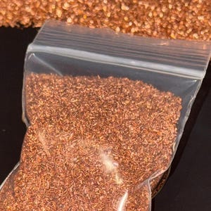May include: A clear plastic bag filled with small, reddish-brown pieces, possibly decorative or craft materials. The bag is sealed and sits on a dark surface, surrounded by scattered pieces of the same material.