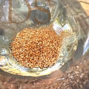 May include: A clear glass container filled with small, irregularly shaped, light brown granules. The granules are clustered together, filling the bottom of the container. The letters "DS" are faintly visible on the glass.