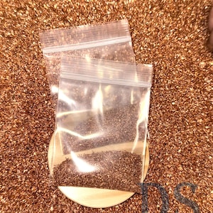 May include: Two clear plastic zip bags filled with small, dark brown particles, resting on a light wooden disc. The background is a textured surface of similar brown particles, creating a cohesive, earthy aesthetic.