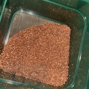 Puede incluir: Un recipiente de plástico verde con una malla fina, parcialmente lleno de material granular de color marrón rojizo. El recipiente tiene esquinas redondeadas y una superficie ligeramente texturizada, para tamizar o filtrar.