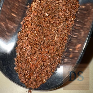 May include: Close-up of a metal scoop filled with dried, reddish-brown tea leaves. The leaves are small, irregularly shaped, and appear to be a blend of different sizes and textures. The scoop is shiny and reflects the light.