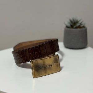 May include: A brown leather belt with a crocodile texture. The belt has a rectangular gold-coloured buckle with the words "DOLCE & GABBANA" engraved on it. The belt is approximately 102 cm long.