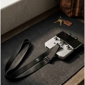 May include: A white drone controller with a black smartphone attached, resting on a dark gray surface. A black lanyard with a logo is connected to the controller. Keys and a brown leather bag are in the background.