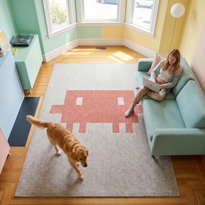 May include: A light gray area rug with a large coral-colored pixelated design in a living room setting. A golden retriever walks across the rug. A person sits on a light blue sofa with a laptop. The room has pastel-colored walls and wooden floors.