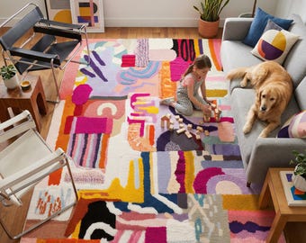 Pode incluir: Um tapete de área abstrato colorido com um design de patchwork. O tapete apresenta uma variedade de formas e cores, incluindo rosa, laranja, amarelo, roxo e branco. Uma criança está brincando no tapete com blocos de madeira, e um cão golden retriever está descansando em um sofá cinza.