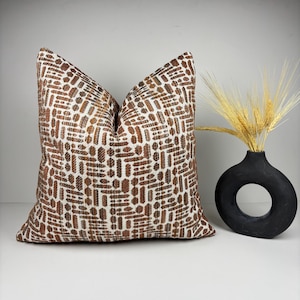 May include: A decorative throw pillow with a brown and white geometric pattern. The pillow is next to a black vase with a circular opening, holding wheat stalks. The pillow's pattern consists of rectangular and circular shapes.