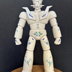 Peut inclure: Figurine de robot blanc avec des motifs floraux bleus et turquoise. La figurine a une tête détaillée, des épaules larges et des membres articulés. Le robot est posé sur une surface en bois sur fond sombre. Le design rappelle un jouet vintage.
