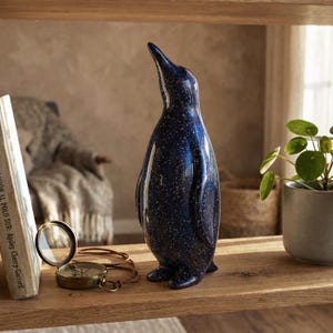 Op de afbeelding: Een staand pinguïnbeeldje met een glanzende donkerblauwe afwerking en een gespikkeld wit patroon dat doet denken aan een sterrenhemel. Het gladde, afgeronde beeldhouwwerk staat rechtop met de snavel lichtjes naar boven en buiten gericht.