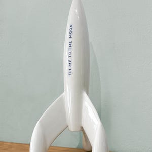 Może przedstawiać: Biała ceramiczna rakieta z ostrym czubkiem i trzema płetwami. Tekst "FLY ME TO THE MOON" jest wydrukowany na boku na niebiesko. Rakieta to przedmiot dekoracyjny.
