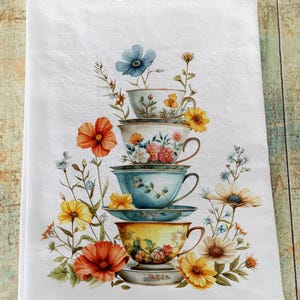 May include: White tea towel featuring a stack of teacups with floral designs and colorful flowers. The teacups are in shades of blue, yellow, and white, with a variety of flowers including orange poppies and yellow daisies. The text "Epoc Ink" is at the top.