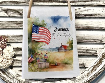 May include: A white tea towel with a watercolor painting of an American flag waving in a field. The text "America the beautiful" is written in script. The towel is held by two wooden clothespins.