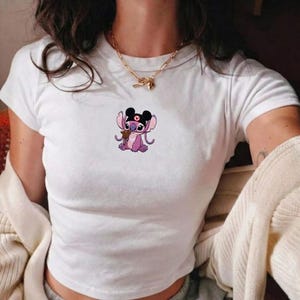 May include: White crop top featuring a cartoon graphic of a purple alien character with black ears and a heart on its head. The character is holding a small brown object. The shirt has short sleeves.