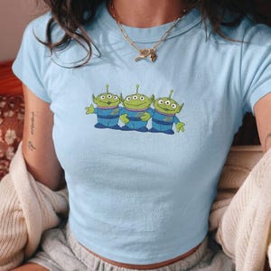 May include: A light blue short-sleeved t-shirt featuring an embroidered graphic of three green three-eyed aliens wearing blue uniforms. The aliens are positioned in a row across the center of the chest, each with a small yellow emblem on their chest.