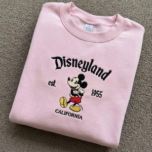 May include: A light pink crewneck sweatshirt featuring black embroidered text that reads Disneyland est. 1955 CALIFORNIA. Centered below the text is an embroidered graphic of Mickey Mouse wearing red shorts and yellow shoes on a light pink fabric.
