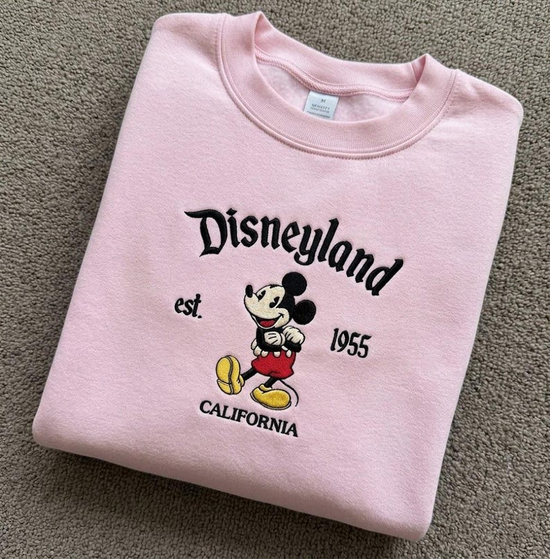 May include: A light pink crewneck sweatshirt featuring black embroidered text that reads Disneyland est. 1955 CALIFORNIA. Centered below the text is an embroidered graphic of Mickey Mouse wearing red shorts and yellow shoes on a light pink fabric.