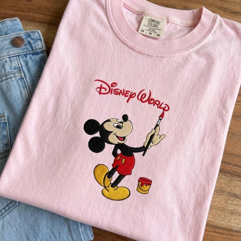 K&ouml;nnte beinhalten: Hellrosa Rundhals-T-Shirt mit gesticktem Mickey Mouse-Design. Mickey wird mit einem Pinsel dargestellt, der die Worte "Disney World" in Rot malt. Das Shirt wird mit einer blauen Jeans kombiniert.