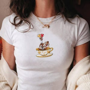May include: White t-shirt featuring an embroidered design of two cartoon chipmunks in a teacup, with balloons. The teacup is yellow with a gold trim. The chipmunks are brown and white.