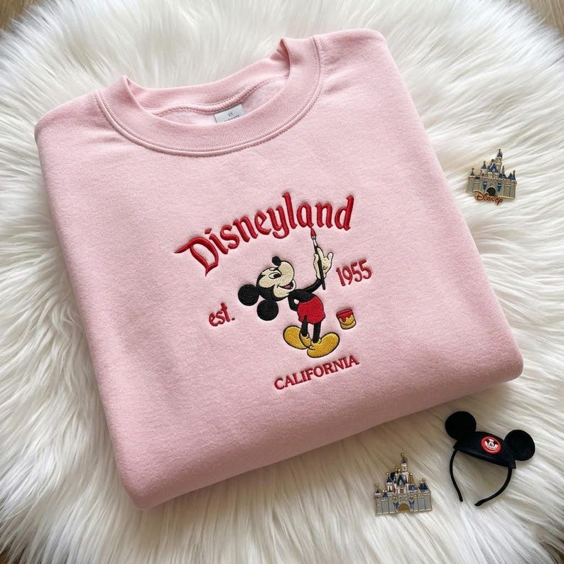 May include: A light pink crewneck sweatshirt featuring an embroidered graphic of Mickey Mouse holding a paintbrush. The text reads Disneyland, est. 1955, and CALIFORNIA in red thread. A small paint can is also embroidered next to the character.