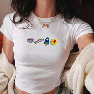 May include: White t-shirt with embroidered cartoon graphics. The design includes the word "ANDY", a purple and green ray gun, a blue rocket ship, and a yellow ball with a red star. The shirt has short sleeves and a crew neck.