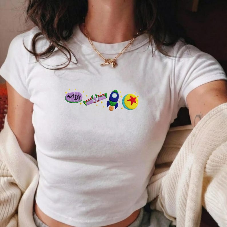 May include: White t-shirt with embroidered cartoon graphics. The design includes the word "ANDY", a purple and green ray gun, a blue rocket ship, and a yellow ball with a red star. The shirt has short sleeves and a crew neck.