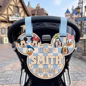 May include: A wooden stroller sign shaped like a Mickey Mouse head with a light blue and white checkered pattern. It features cartoon character faces, the text "the SMITH family", and small star and castle icons, attached by two light blue straps.