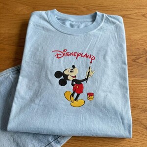 Pu&ograve; includere: T-shirt azzurra con logo Disneyland rosso e illustrazione di Topolino con un pennello. Topolino indossa pantaloncini rossi e scarpe gialle. &Egrave; visibile anche un piccolo barattolo di vernice. La maglietta &egrave; piegata su una superficie di legno.