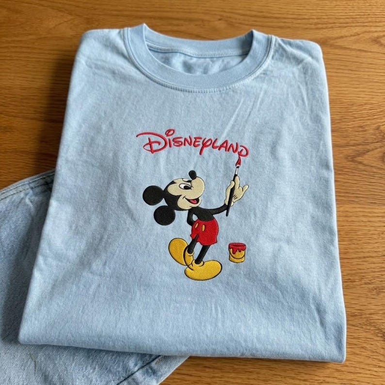 Pu&ograve; includere: T-shirt azzurra con logo Disneyland rosso e illustrazione di Topolino con un pennello. Topolino indossa pantaloncini rossi e scarpe gialle. &Egrave; visibile anche un piccolo barattolo di vernice. La maglietta &egrave; piegata su una superficie di legno.