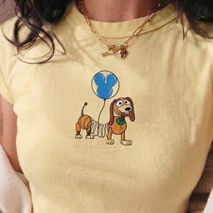 Op de afbeelding: Lichtgeel T-shirt met een geborduurd cartoonhondje dat een blauwe ballon vasthoudt in de vorm van een Mickey Mouse-hoofd. De hond heeft bruin en beige haar en een groene kraag.