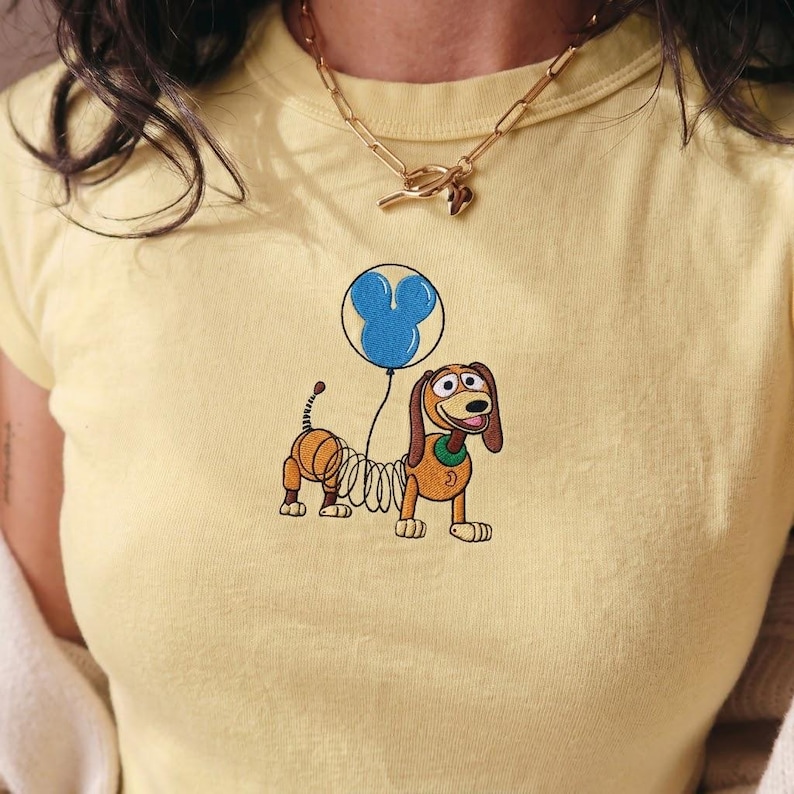 Op de afbeelding: Lichtgeel T-shirt met een geborduurd cartoonhondje dat een blauwe ballon vasthoudt in de vorm van een Mickey Mouse-hoofd. De hond heeft bruin en beige haar en een groene kraag.