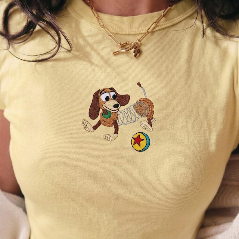 May include: A light yellow t-shirt featuring an embroidered cartoon dog with a spring body, a brown ear, and a green collar. The dog is playing with a red star and yellow ball. A gold necklace is worn.