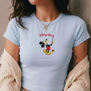 Pu&ograve; includere: T-shirt azzurra con una grafica di Topolino che dipinge, con le parole "Disney World" in rosso. Topolino &egrave; raffigurato nel suo abito classico, con un pennello e accanto a un barattolo di vernice. La maglietta ha maniche corte.
