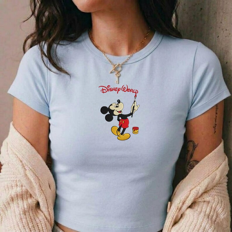 Pu&ograve; includere: T-shirt azzurra con una grafica di Topolino che dipinge, con le parole "Disney World" in rosso. Topolino &egrave; raffigurato nel suo abito classico, con un pennello e accanto a un barattolo di vernice. La maglietta ha maniche corte.