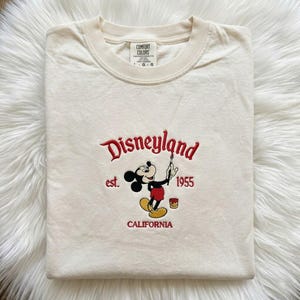 May include: A cream-coloured short-sleeve t-shirt featuring an embroidered design of Mickey Mouse holding a paintbrush. The text reads Disneyland, est. 1955, and CALIFORNIA in red thread. The shirt has a Comfort Colors brand label at the inner neckline.